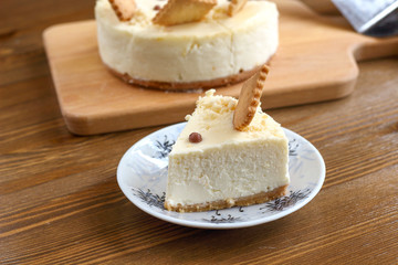 A piece of classic New York cheesecake decorated with square cookies on plate, on wooden table in coffee pastry shop with whole cake in the background. Cake sweet dessert for holiday, birthday party