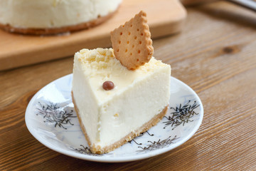 A piece of classic New York cheesecake decorated with square cookies on plate, on wooden table in coffee pastry shop with whole cake in the background. Cake sweet dessert for holiday, birthday party