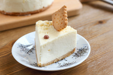 A piece of classic New York cheesecake decorated with square cookies on plate, on wooden table in coffee pastry shop with whole cake in the background. Cake sweet dessert for holiday, birthday party