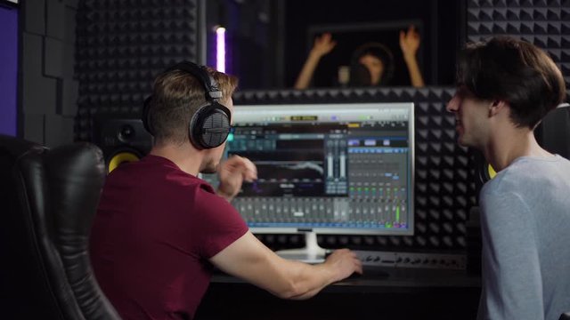 Back view tracking shot of sound engineer and producer using computer with professional software and discussing details while recording song of young Asian woman in recording studio