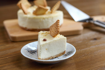 A piece of classic New York cheesecake decorated with square cookies on plate, on wooden table in coffee pastry shop with whole cake in the background. Cake sweet dessert for holiday, birthday party