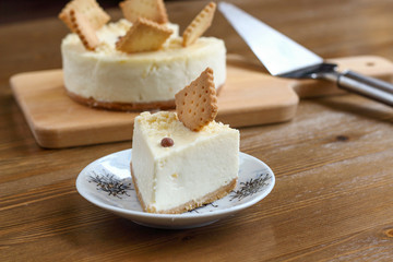 A piece of classic New York cheesecake decorated with square cookies on plate, on wooden table in coffee pastry shop with whole cake in the background. Cake sweet dessert for holiday, birthday party