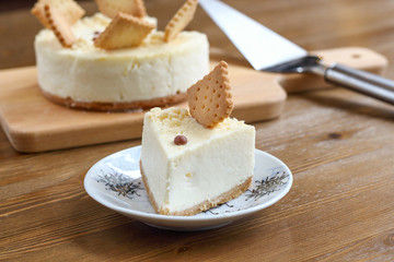 A piece of classic New York cheesecake decorated with square cookies on plate, on wooden table in coffee pastry shop with whole cake in the background. Cake sweet dessert for holiday, birthday party