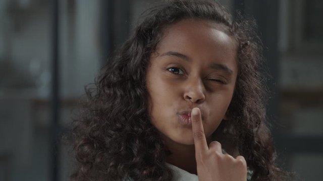 Portrait of charming preadolescent african american girl with curly hair holding index finger on lips and blowing kiss with playful and mischievous expression, looking with beaming smile indoors.