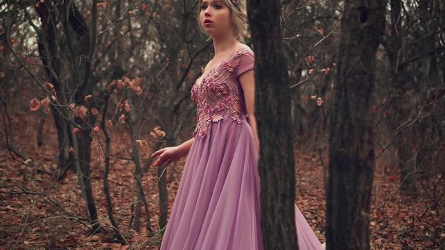 beauty blonde princess is standing in purple lush dress in a corset. Frightened, lost in the forest. Glamorous fashion woman queen in crown. Autumn backdrop, black trees orange leaves. Cute young face
