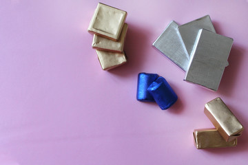 Pile of chocolates wrapped in silver, golden and blue foil. Copy space for text. Template for greeting card, poster or invitation. Sweet candies on pink smooth table. Backdrop or background