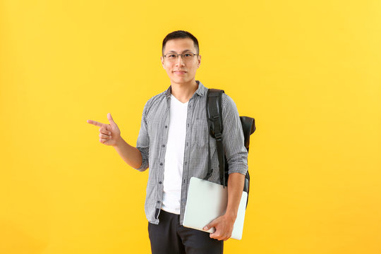 Male Asian student with laptop pointing at something on color background