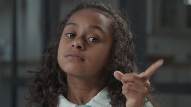 Portrait of lovely strict preadolescent black girl with curly hair showing no, don't, forbidden, warning with finger hand gesture, making reprimand or rebuke indoors, looking with determination.