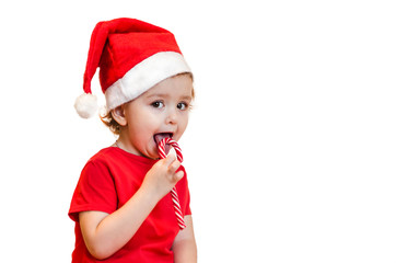 the baby in a santa hat and red dress eats and licks a candy cane with appetite. Christmas sweets and gifts for children. new year conception. copy space, layout, banner, postcard
