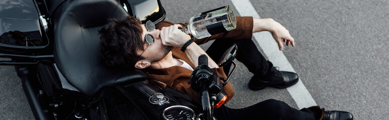 panoramic shot of young motorcyclist sitting on ground, leaning on motorcycle and drinking alcohol
