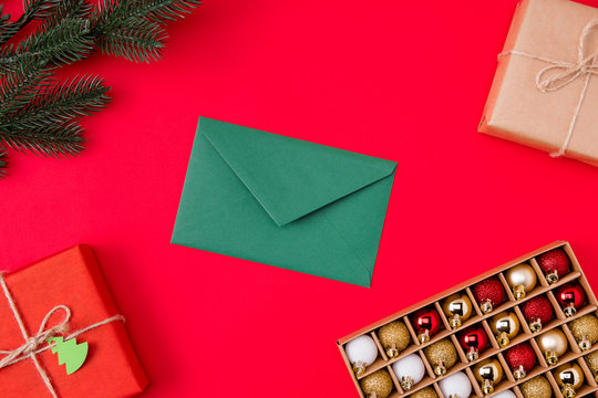 Top above high angle view photo of green envelope surrounded with gifts presents branch of fir tree and box of christmas toys isolated vibrant color red background