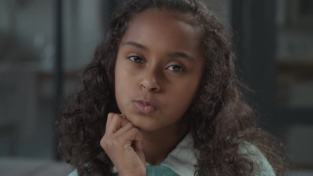 Pensive adorable preteen african girl in deep thoughts propping up chin with hand, contemplating about something, making choice, looking puzzled, with placid and thoughtful look indoors.