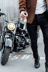 cropped view of man in brown jacket walking near motorcycle and holding alcohol bottle