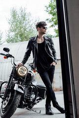 Fototapeta na wymiar full length view of handsome man in leather jacket standing near motorcycle and leaning on it
