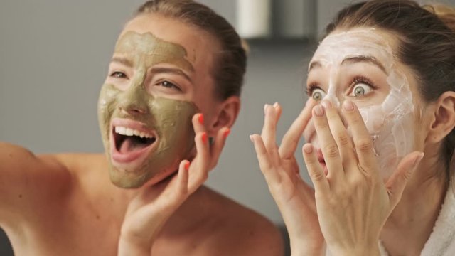 Two pretty smiling girls friends with facial mask on their faces posing and enjoying the moment at home