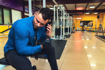 Handsome young man in blue shirt and tie, talking on the phone with a dumbbell in hand in the gym