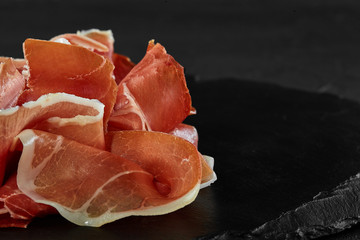 Gourmet, manually sliced jamon on black stone slate board against a dark grey background. Close-up shot. Side view. Copy space.