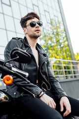 Obraz na płótnie Canvas serious motorcyclist in sunglasses and black leather jacket sitting on motorcycle and looking away
