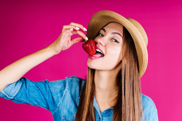 girl with a lovely smile in a hat playfully brings the berry to her mouth in a pink studio