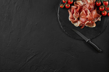 Sliced jamon, cherry tomatoes and a knife on black stone slate board against a dark grey background. Close-up shot. Top view. Copy space.