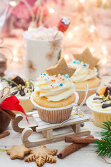 Christmas decorated cupcakes on wooden sledge. Cinnamon rolls, cotton flowers, tangerine bites, christmas tree branches and gingerbread cookies. String lights and cup with marshmallow on background.