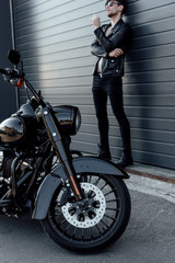 Obraz na płótnie Canvas full length view of motorcyclist in sunglasses and leather jacket standing near wall close to motorcycle and holding cigarette