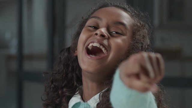 Portrait of excited lovely preteen african girl with curly hair making hand gun gesture, pointing forward and laughing out loud, expressing joy, carefree playful mood, happiness and positivity.