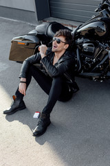 Fototapeta na wymiar handsome motorcyclist in leather jacket smoking cigarette and smiling while sitting near motorcycle