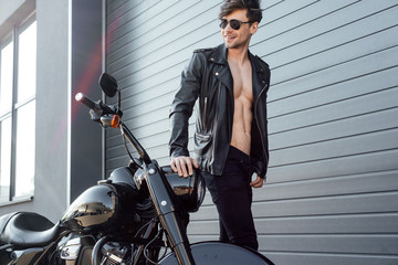 Fototapeta na wymiar young man in sunglasses with leather jacket and naked torso standing near black motorcycle, smiling and looking away
