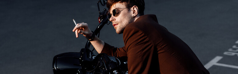 panoramic shot of young man sitting on motorcycle and smoking cigarette