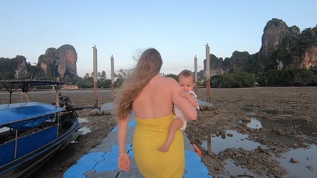A woman in a yellow dress carries in her arms a child on a pontoon pier during a high tide. Mom walks with her daughter at dawn at sea. Family holiday.