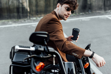 selective focus of handsome man in sunglasses and brown jacket sitting on motorcycle and holding coffee to go