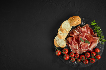 Sliced jamon, cherry tomatoes, baguette, herbs and spices on black stone slate board against a dark...