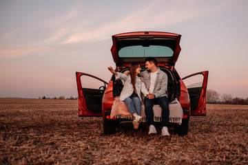 Young Happy Couple Dressed Alike in White Shirt and Jeans Sitting at Their New Car Trunk, Beautiful Sunset on the Field, Vacation and Travel Concept