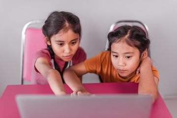 The older sister, who is wearing a pink T-shirt, is using internet for searching knowledge on laptop with young sister who is wearing an orange T-shirt. Both of them braid and cute white ribbons.