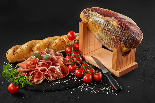 Jamon carved from pig leg on jamonera stand, cherry tomatoes, herbs on black stone slate board, baguette and knife on dark background. Close-up.