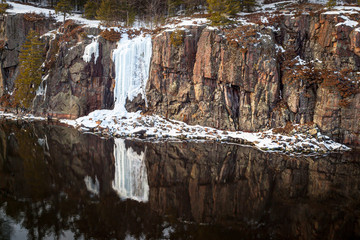A white frozen waterfall along a dark brown sheer cliff of rock alone a placid river