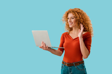 smiling girl waving and having video call on laptop isolated on blue