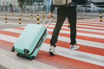 Picture of man walking on crosswalk and carrying blue suitcase with packpack on back. Going to airport or railway station alone. Cut view. Vacation or trip.