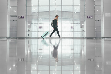Side view of lonely single man walking in airport hall. Gong to business trip or vacation. Travel with blue suitcase and backpack. Floor with reflection. Daylight.