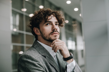 Side view of young modern businessman stand in room and think. Calm peaceful guy look up and hold hand under chin. Expert or diplomat. Posing on camera. Wear official suit or jacket.