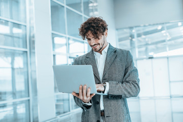 Fototapeta premium Blue picture of young businessman with laptop in hands typing on keyboard. Stand in room and smile. Confident expert on picture. Work remote alone. One in room. Modern technologies.