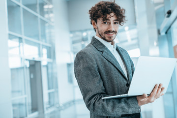 Young cheerful businessman stand in room and smile. Posing with laptop in hands. Work remote. Happy...