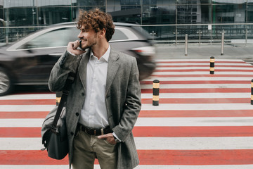 Cheerful young businessman in suit stand on crosswalk alone and talk on phone. Hold one hand in...