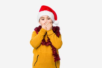Little boy celebrating christmas day wearing a santa hat isolated shocked covering mouth with hands.