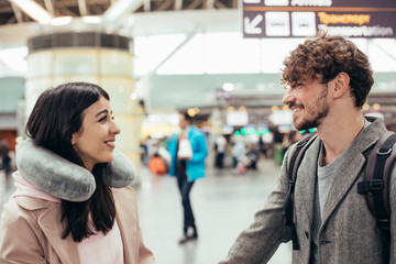 Cheerful attractive beautiful couple in airport. Young man and woman look at each other and smile. Ready to travel abroad. Waiting for flight. Trip or vacation. Travel pillow around her neck.