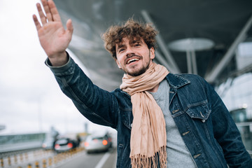 Portrait of young man showing palm of his hand. Trying to catch taxi cab or transportation. Look straight ahead. Stand at airport alone. Daylight. After travel or trip abroad. © Anton