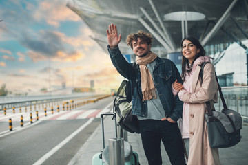 Young man and woman stand outside airport and wait for taxi cab. Guy wave with hand. After vacation or travel. Georgian woman hold his hand and smile. Sunset outside. Evening.