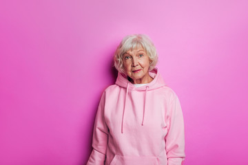 Amazing old woman in pink sweater looks modern and good-looking. Stand alone and look forward. Calm, peaceful and confident senior female model in hoody. Isolated over pink background.