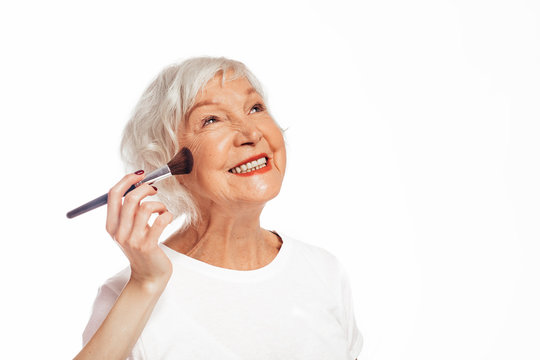 Happy, cheerful and positive old woman smiling. Stand alone and wait while young hand put some make-up on with brush. Peparing. Isolated over white background.